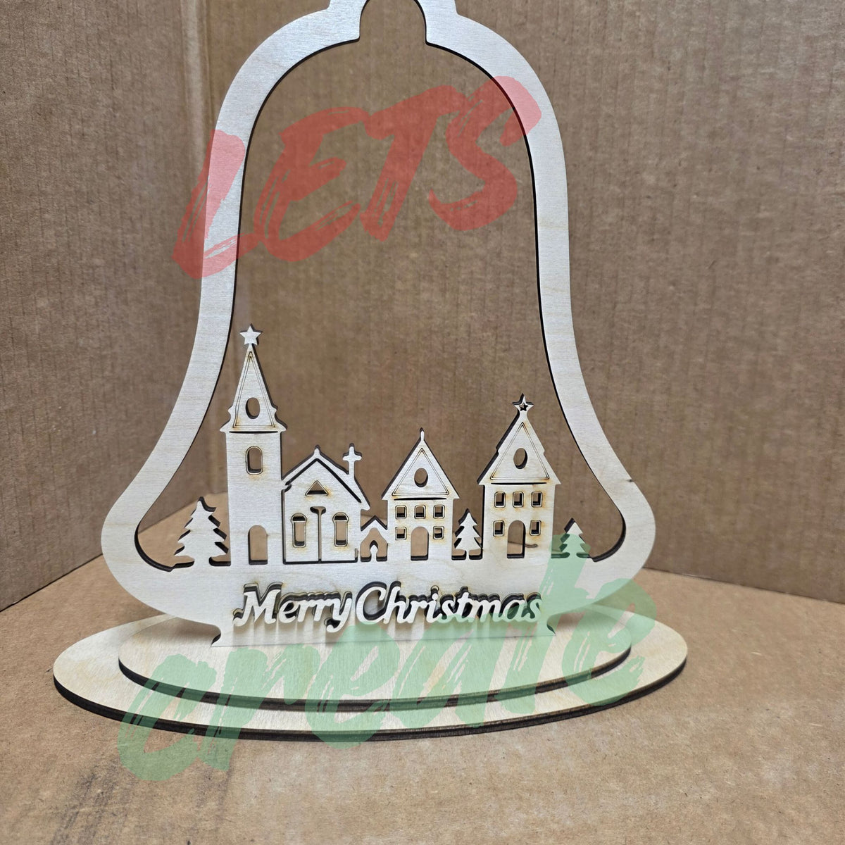 A bell-shaped wooden Christmas ornament featuring a layered nativity scene with a church, houses, pine trees, and a star, with the script words "Merry Christmas" displayed against a neutral background.