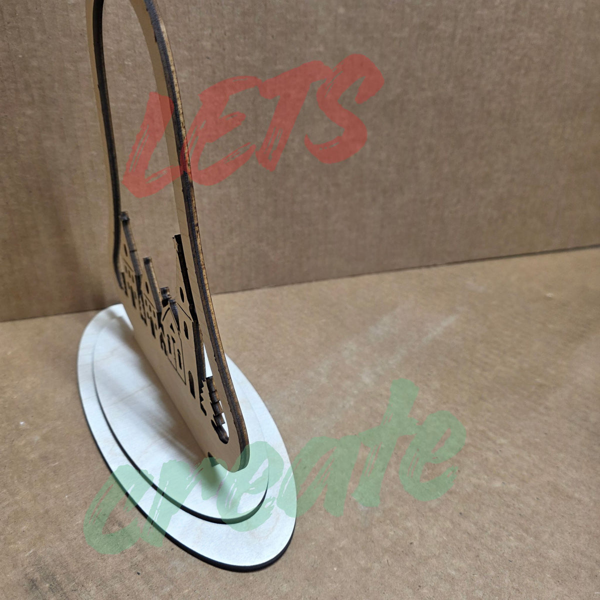 A bell-shaped wooden Christmas ornament featuring a layered nativity scene with a church, houses, pine trees, and a star, with the script words "Merry Christmas" displayed against a neutral background.