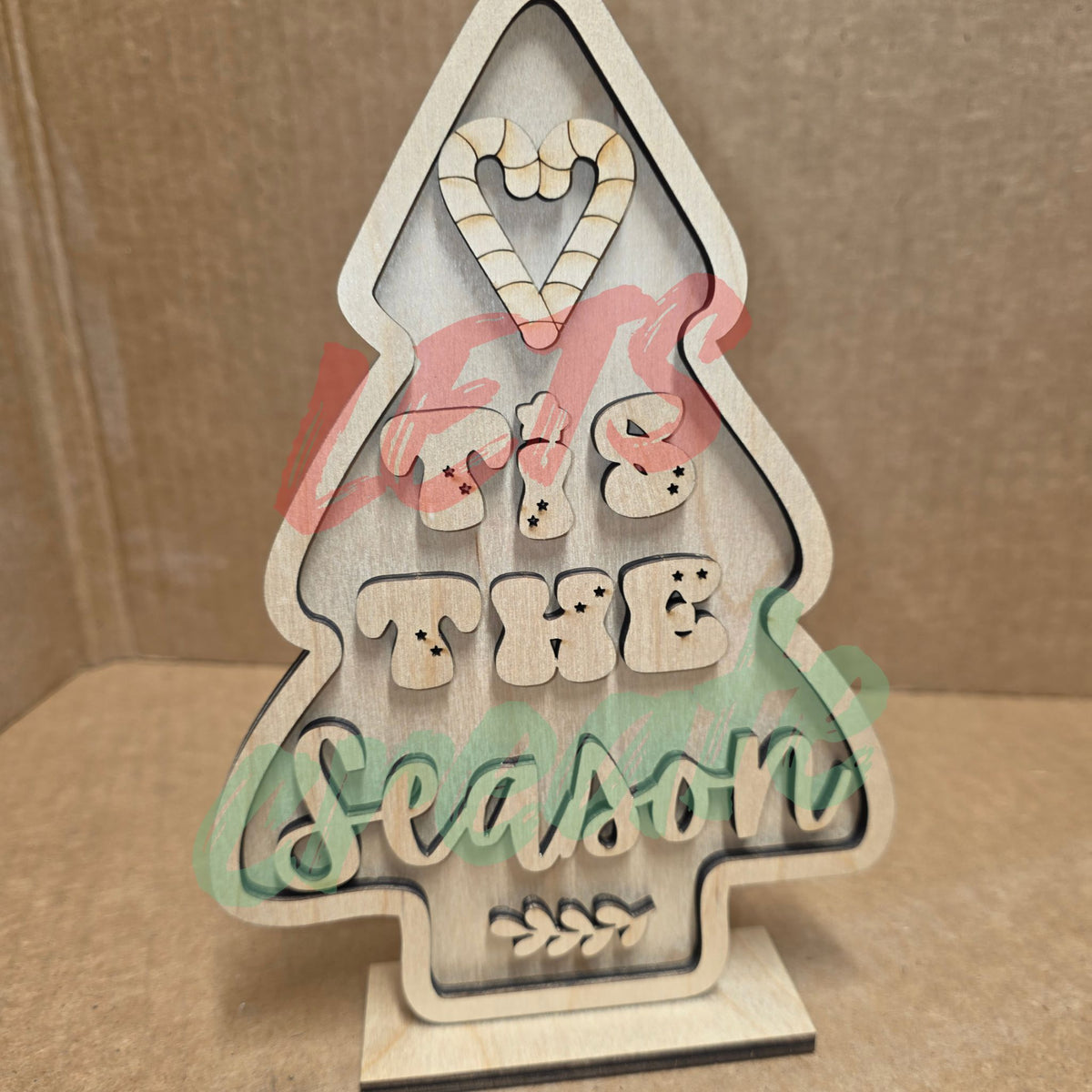 A layered wooden Christmas tree sitter sign featuring a tree-shaped design with candy-cane-striped accents, candy elements, and the script words "Tis the Season" displayed against a neutral background.