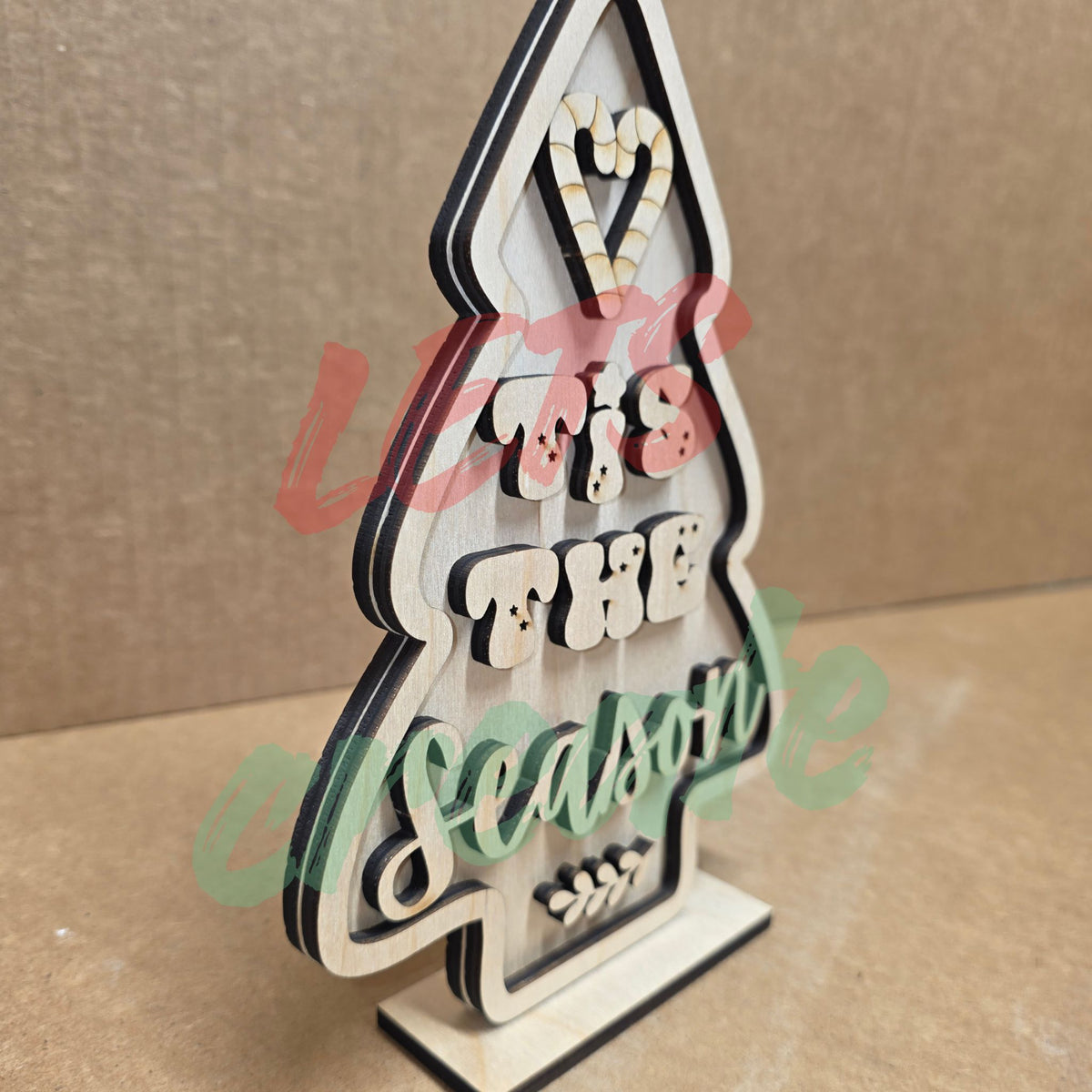 A layered wooden Christmas tree sitter sign featuring a tree-shaped design with candy-cane-striped accents, candy elements, and the script words "Tis the Season" displayed against a neutral background.
